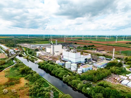Aerial view of an industrial power plant near a river, surrounded by greenery and wind turbines on a cloudy day.